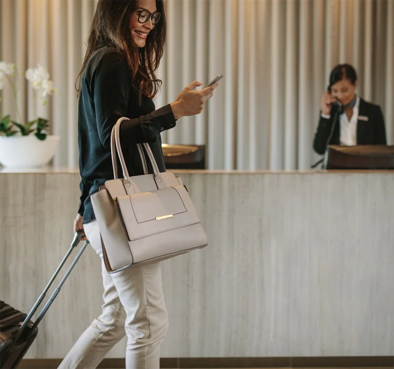 Woman with luggage walking through hotel lobby checking phone while receptionist works at desk - in Chevy Chase, MD