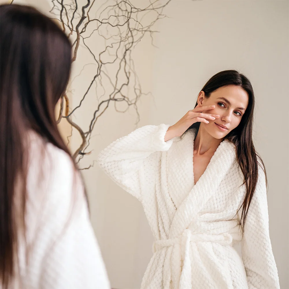 Woman in white bathrobe applying skincare product while looking in mirror during morning beauty routine - Skin rejuvenation in Chevy Chase, MD
