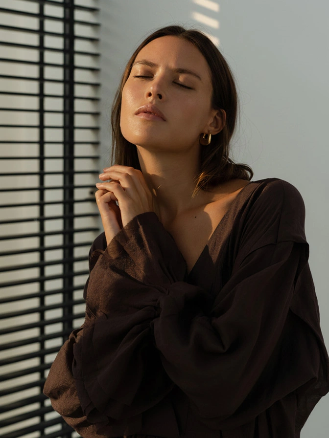 Woman in black blazer with eyes closed standing by window blinds with dramatic sunlight shadows - Clear and brilliant in Chevy Chase, MD
