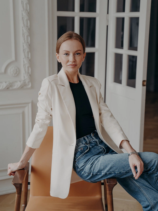 Professional woman in white blazer and jeans sitting on wooden stool in elegant interior setting - Deep plane facelift in Chevy Chase, MD