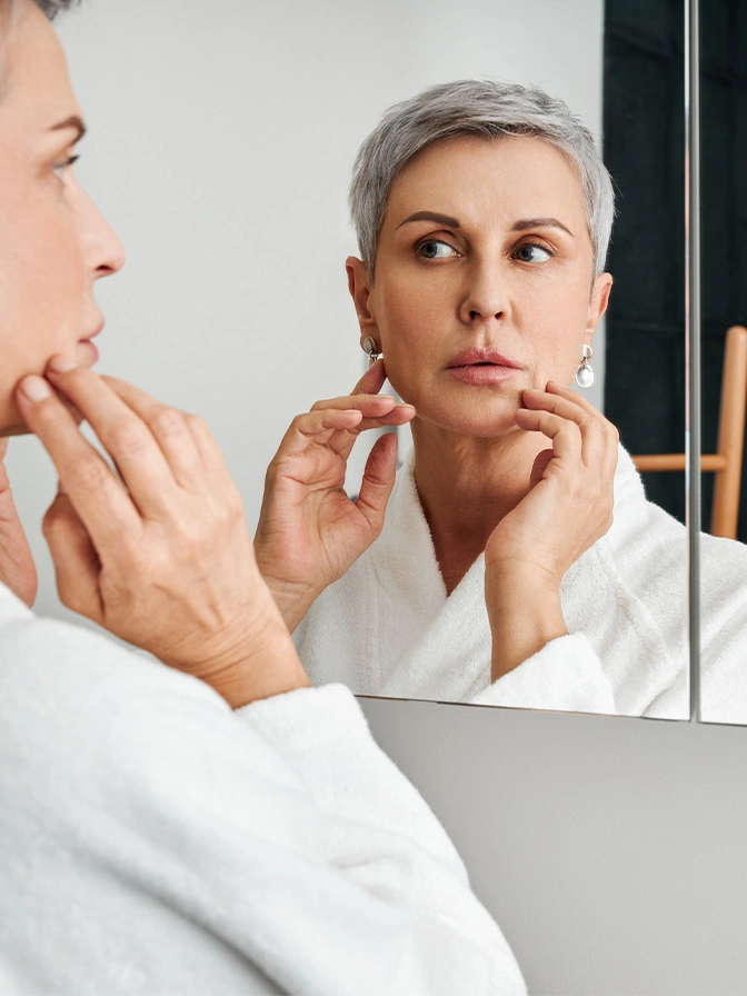 Woman with short gray hair examining her face in bathroom mirror while wearing white bathrobe - Earlobe repair in Chevy Chase, MD