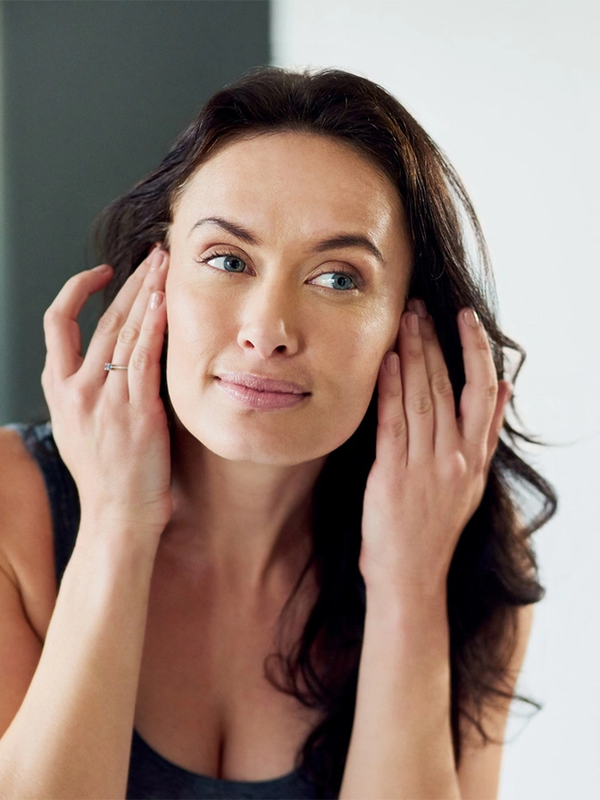 Woman examining her facial skin while touching her face with both hands in mirror - Eyelid surgery in Chevy Chase, MD
