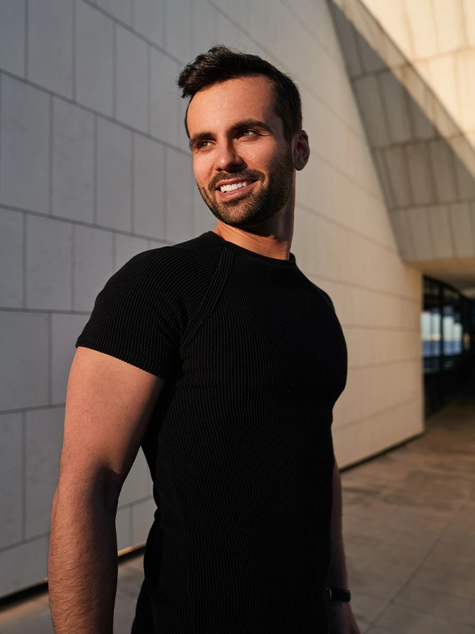Smiling man in black t-shirt standing against modern building wall in natural sunlight - Gynecomastia in Chevy Chase, MD