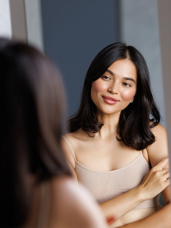 Woman with long dark hair in beige top smiling at her reflection in mirror - IPL photofacial in Chevy Chase, MD