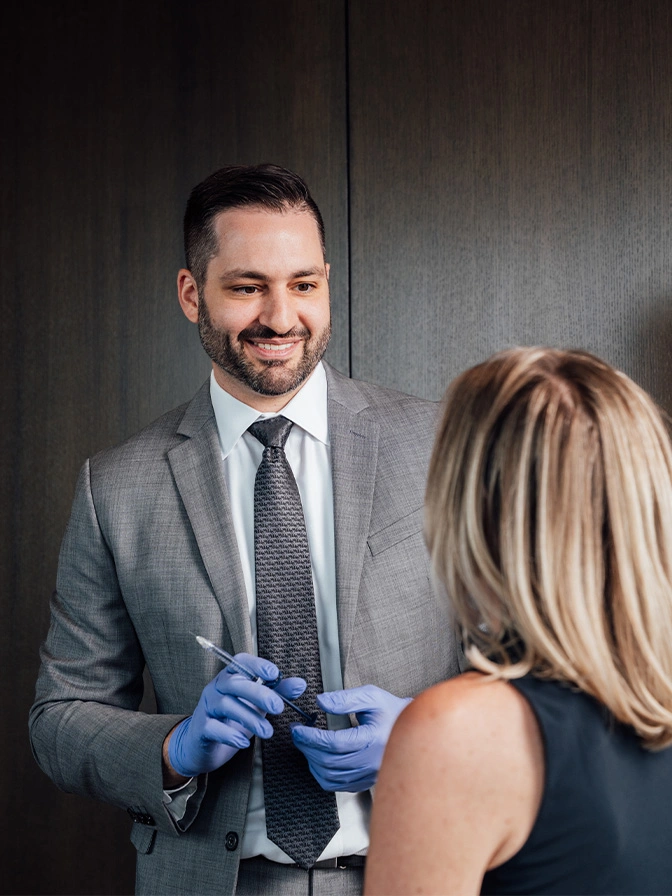 Dr James Economides in gray suit and blue gloves consulting with female patient in modern office - Liquid facelift in Chevy Chase, MD