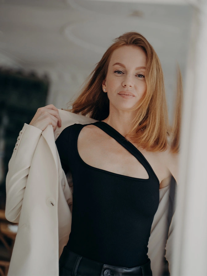 Professional woman with red hair in black dress holding cream blazer, smiling at camera in modern office setting - Nipple reduction in Chevy Chase, MD
