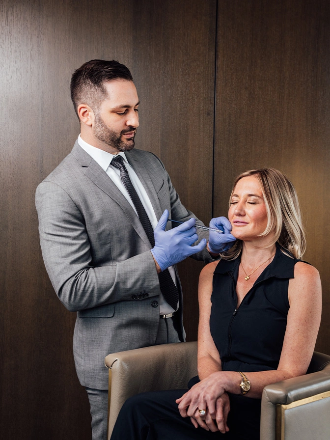 Dr James Economides in grey suit examining female patient's face while wearing blue gloves in consultation room - Non invasive facial balancing in Chevy Chase, MD