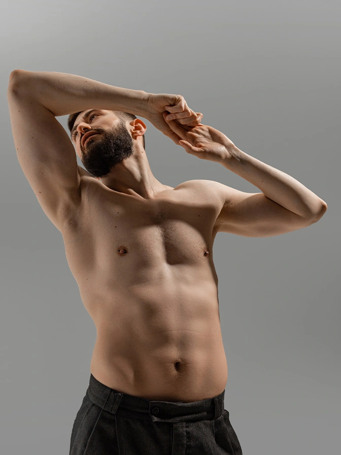 Fit shirtless man stretching arms overhead showing defined abs and muscular torso against gray background - Pec augmentation in Chevy Chase, MD