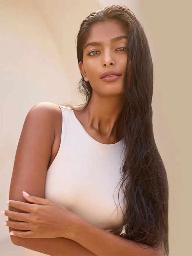 Woman with long dark hair wearing white sleeveless top against neutral background in natural lighting - Preservation breast augmentation in Chevy Chase, MD