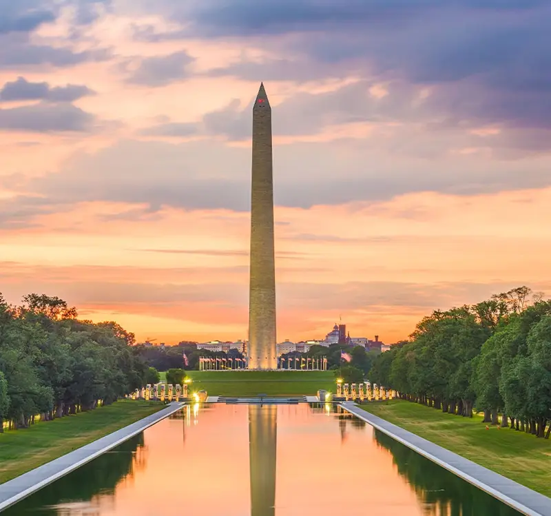 Washington monument at sunset with light reflecting on the pool and very green grass and trees.